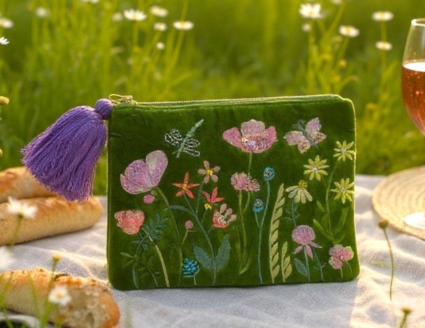 Green embroidered pouch with floral designs on a picnic blanket in a field of flowers.