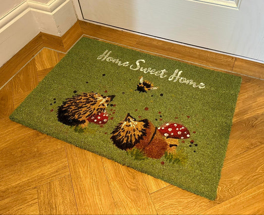 green doormat with script reading home sweet home, images of two hedgehogs and mushrooms