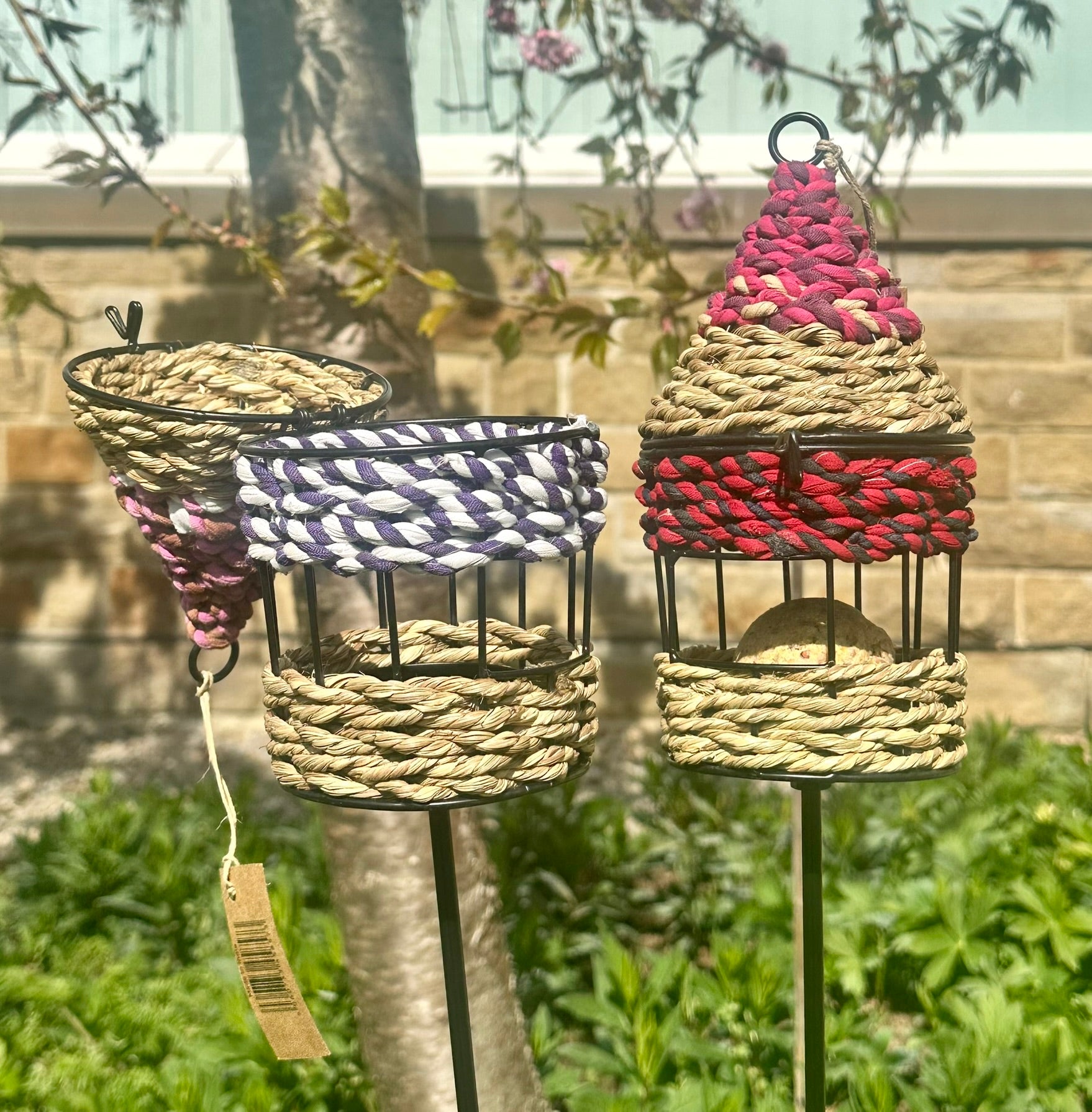 two of the colourful handwoven bird feeders made of munja grass, displayed on garden stakes in a backyard setting