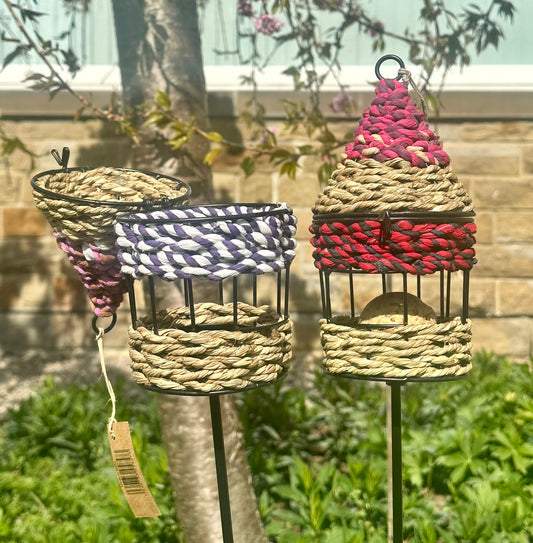 two of the colourful handwoven bird feeders made of munja grass, displayed on garden stakes in a backyard setting