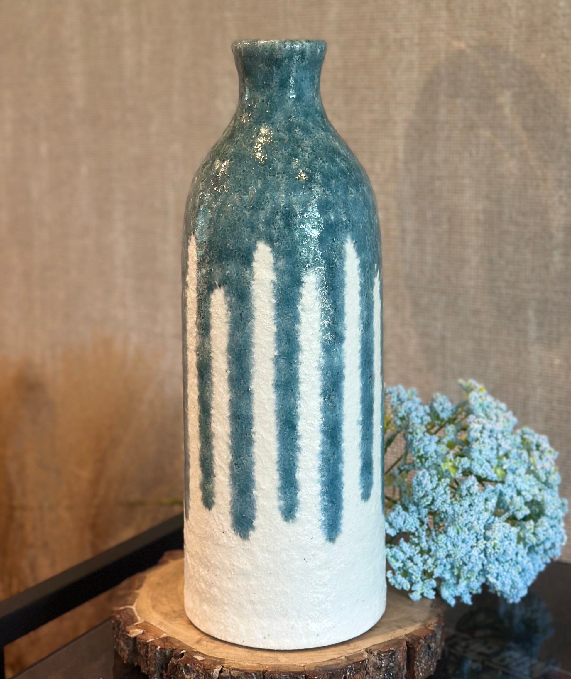 A ceramic bottle vase with a textured blue-green glaze on the upper half and dripping vertical white bands on the lower half, displayed on a rustic wood slice with dried flowers against a neutral fabric backdrop.
