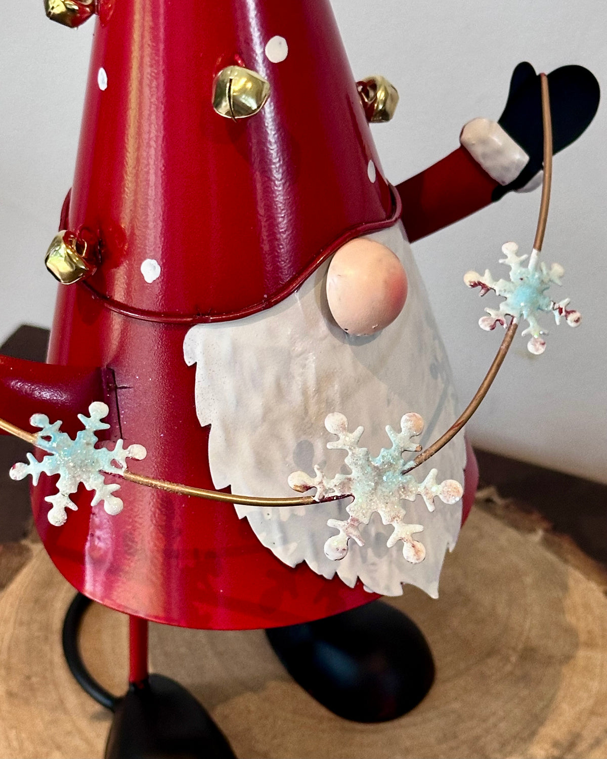 A red metal Christmas gnome decoration with a white beard, black boots, gold bells, and a gold hoop holding white and blue snowflakes, photographed indoors on a wood slice surface.