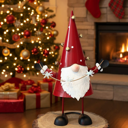 A festive metal Christmas gnome decoration standing on a round wood slice base. The gnome has a tall red cone-shaped hat with small gold bells, a white beard, black boots and gloves, and holds a gold garland with white snowflakes.