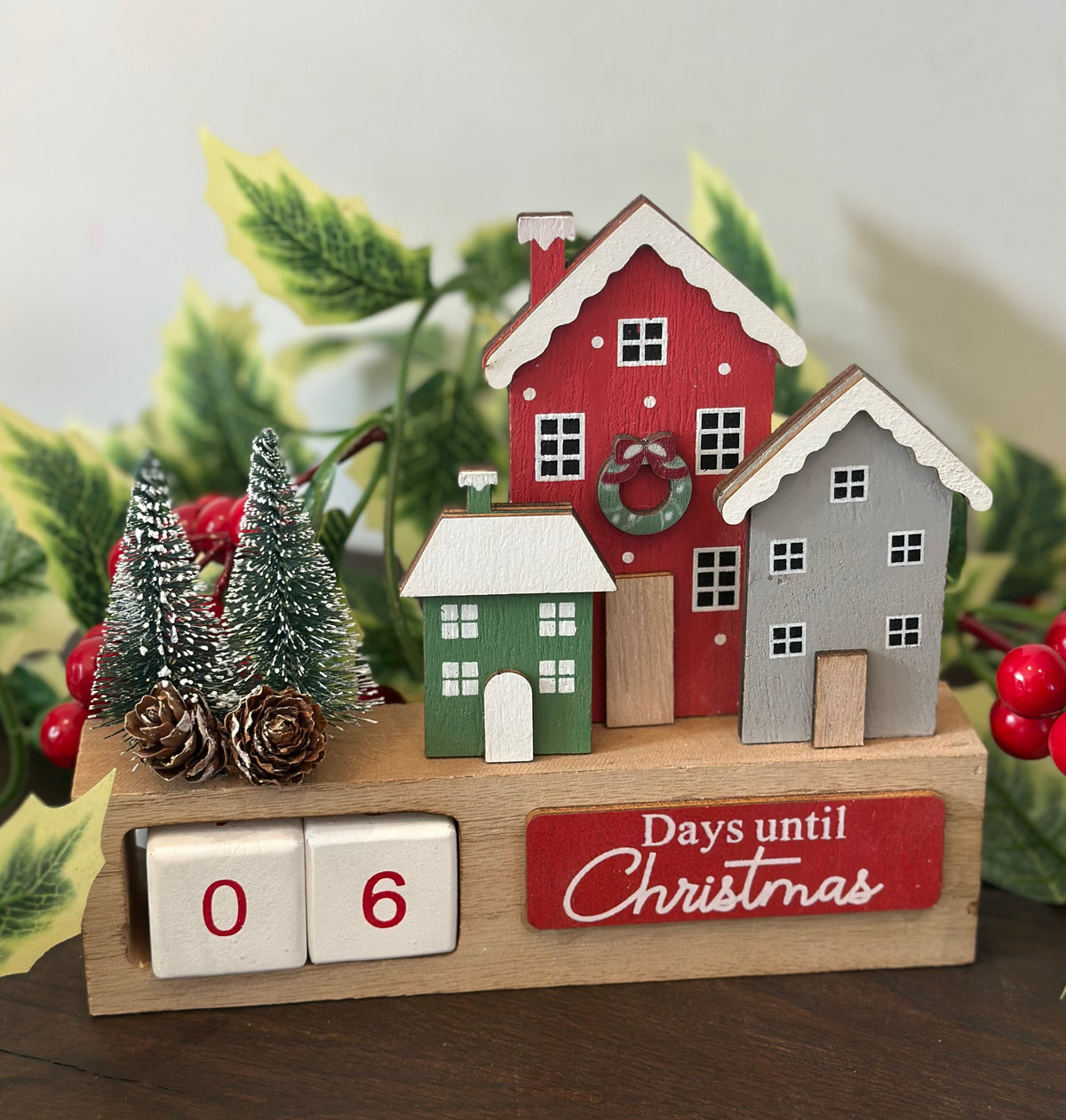 A wooden Christmas countdown calendar display on a tabletop, featuring three small painted houses in red, green, and gray, two miniature trees, pinecones, red berries, and a wooden cube flipper showing 06 days until Christmas. Decorative greenery and red berries frame the display.