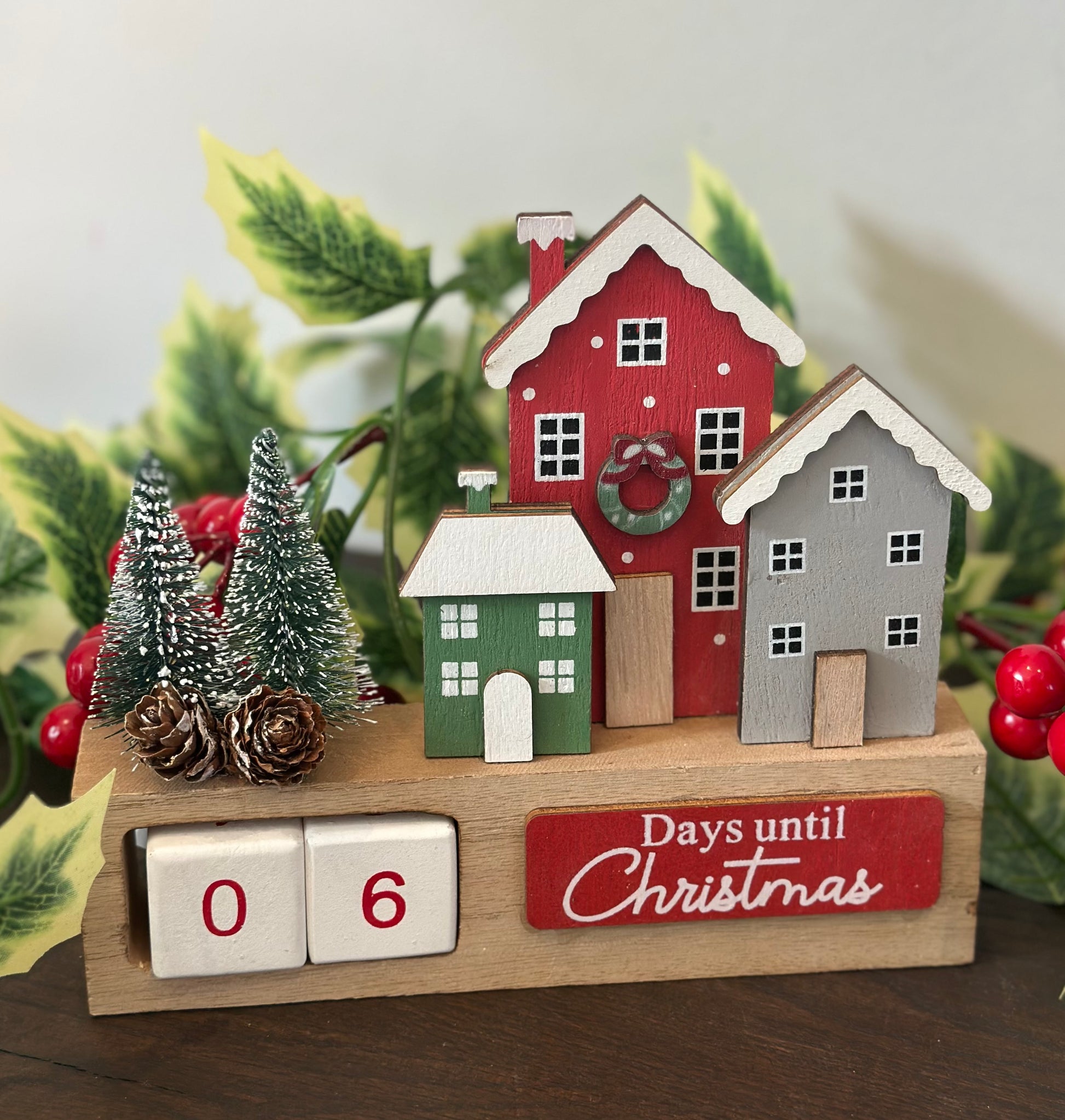 A wooden Christmas countdown calendar display on a tabletop, featuring three small painted houses in red, green, and gray, two miniature trees, pinecones, red berries, and a wooden cube flipper showing 06 days until Christmas. Decorative greenery and red berries frame the display.