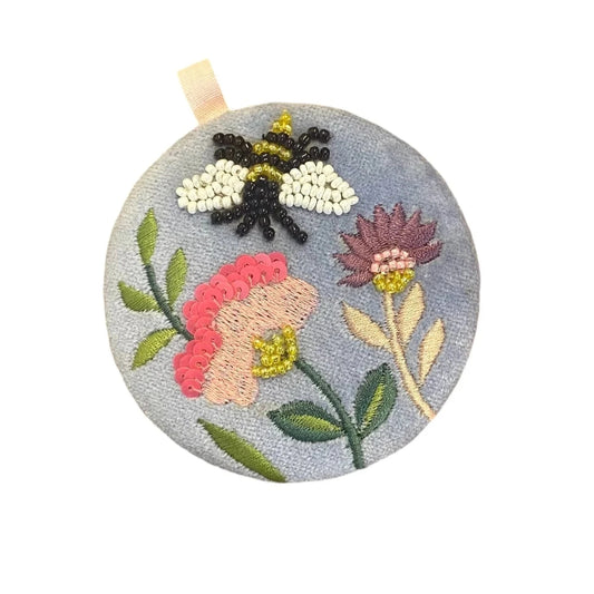 A round embroidered mirror with floral patterns and a beaded bee on a blue background