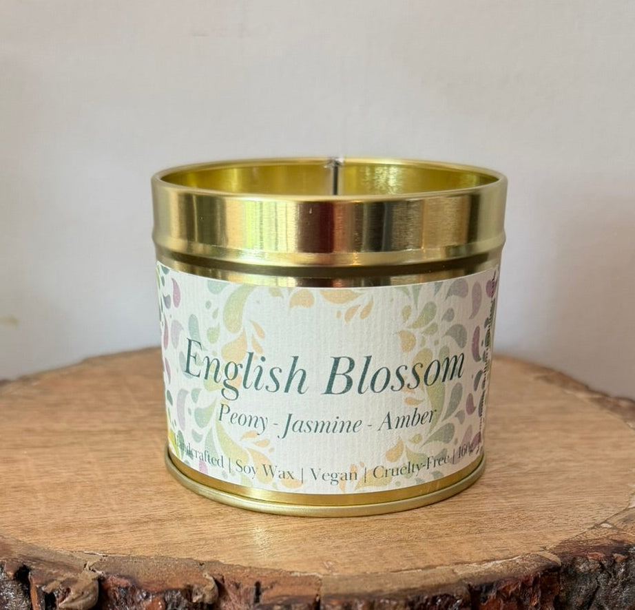 A small candle in a gold metal tin with a white floral label reading 'English Blossom' and 'Peony Jasmine Amber,' displayed on a round natural wood slice coaster over a dark wooden surface.