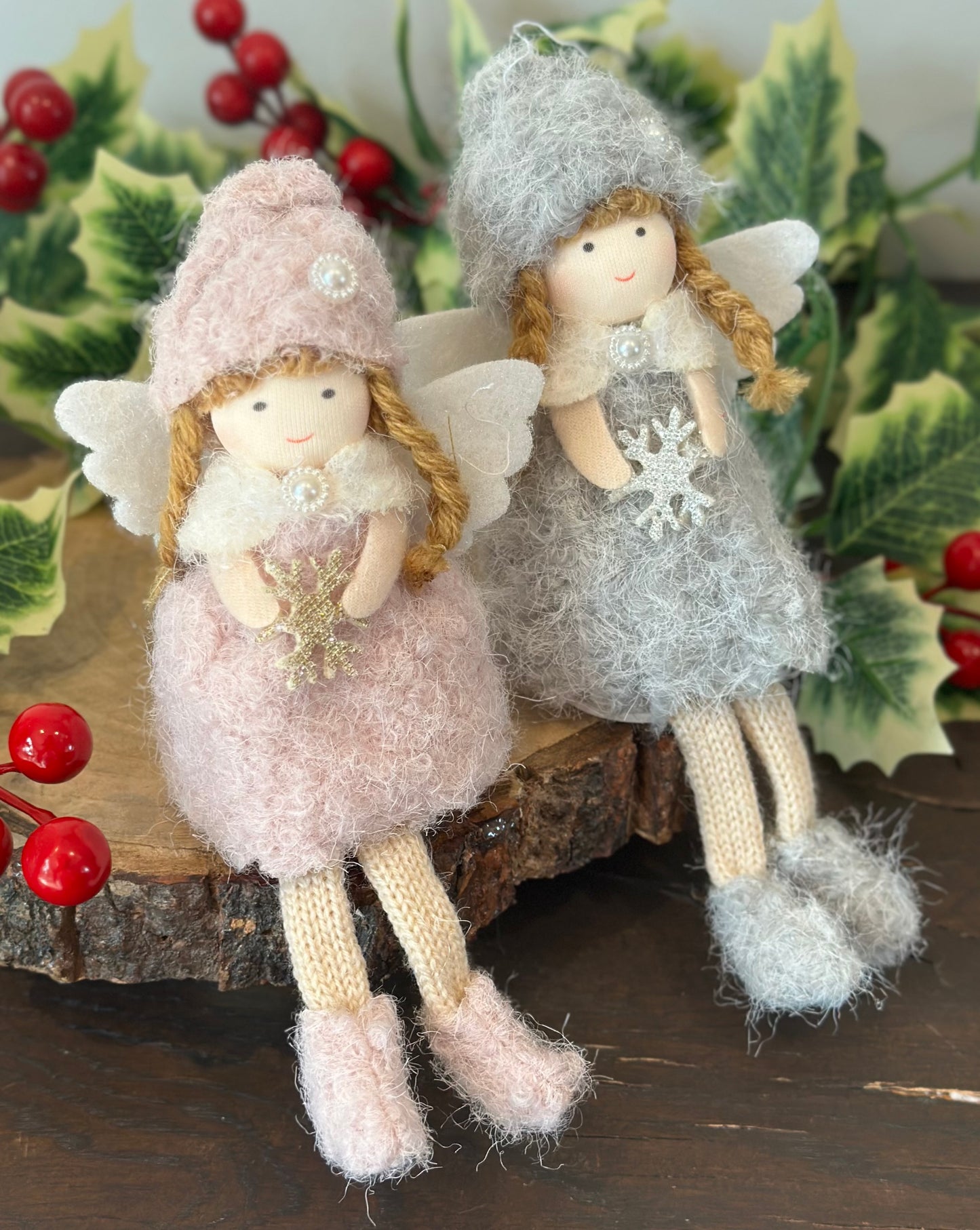 Two handmade wool angel ornaments with white wings, one in dusty pink and one in light gray, sitting on a rustic wood slice with holly and red berries in the background.