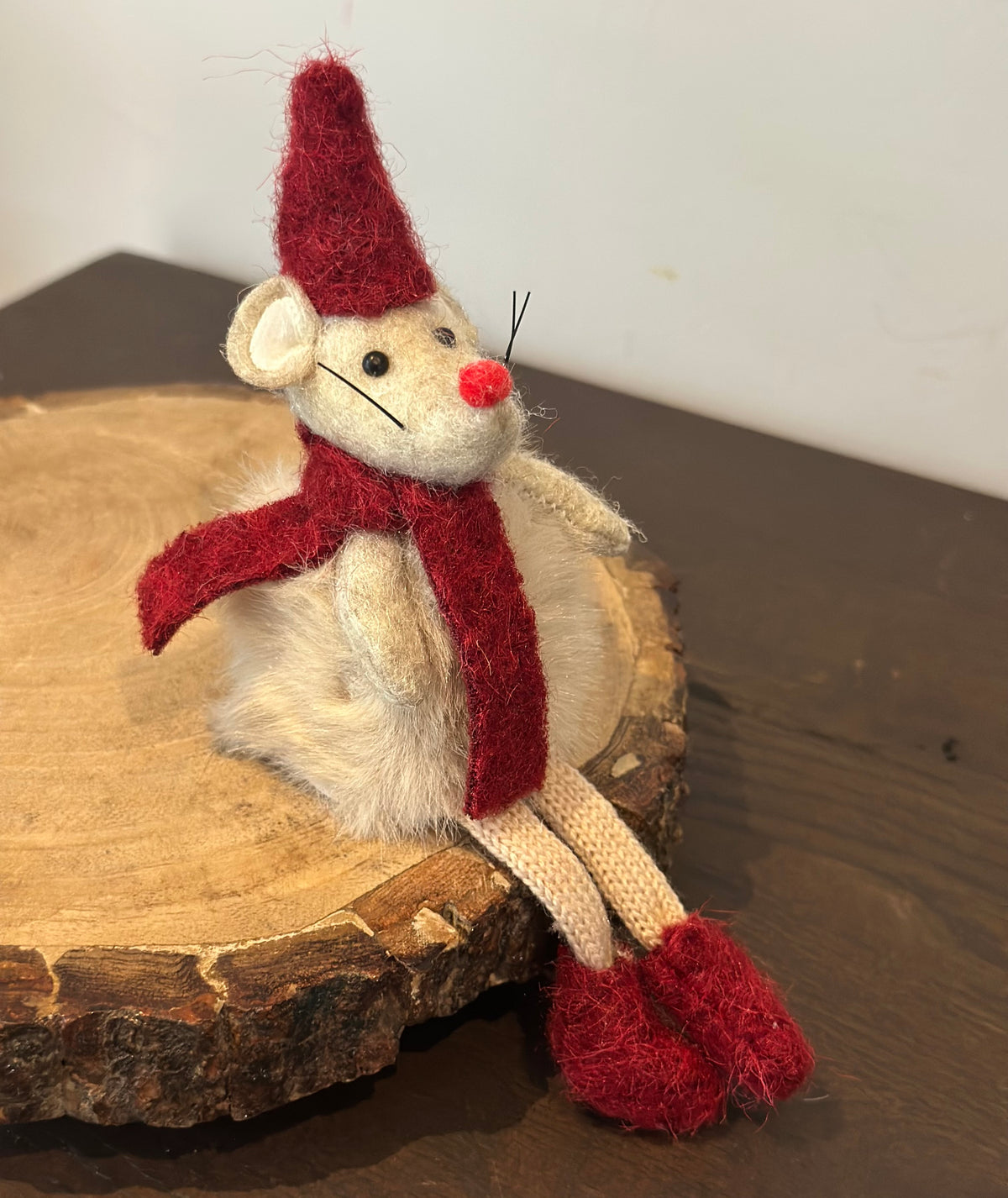 A small felted wool mouse decoration wearing a red pointed hat, red scarf, cream-colored legs, and a red nose, sitting on a rustic wood slice.