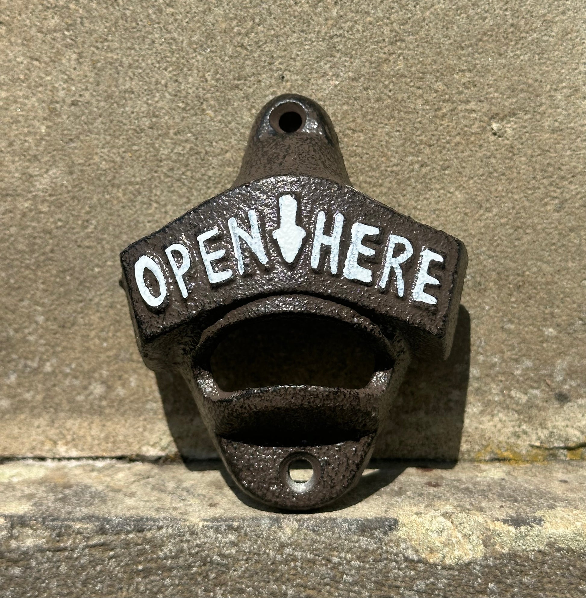 Cast iron wall-mounted bottle opener with 'Open Here' text on a concrete background