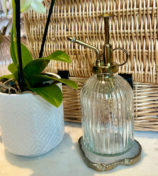 A clear ribbed glass plant mister with a sleek gold finish, positioned next to a wicker basket and a potted plant.