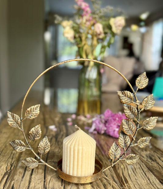 A cream-colored ribbed candle in a round gold-toned holder, surrounded by a decorative gold leaf arch on a rustic wooden table with a blurred background of flowers and a living room.