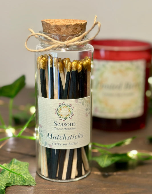 Jar of matchsticks with a cork lid on a wooden surface, surrounded by greenery and a blurred candle.