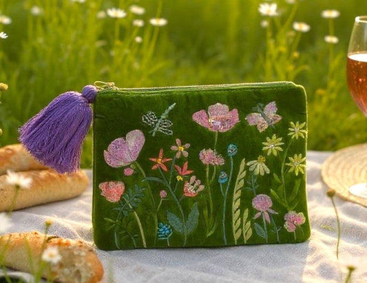 Green embroidered pouch with floral designs on a picnic blanket in a field of flowers.