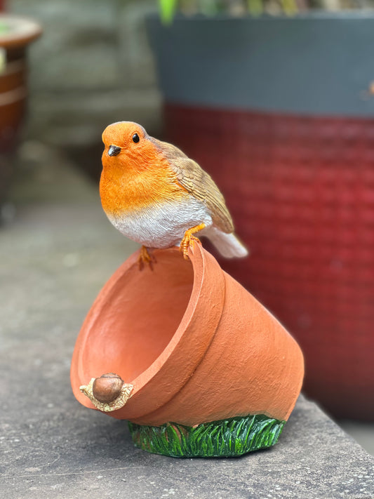A small decorative bird figurine perched on an upside-down terracotta pot with a green grass-like base, photographed outdoors on a stone surface with blurred potted plants in the background.