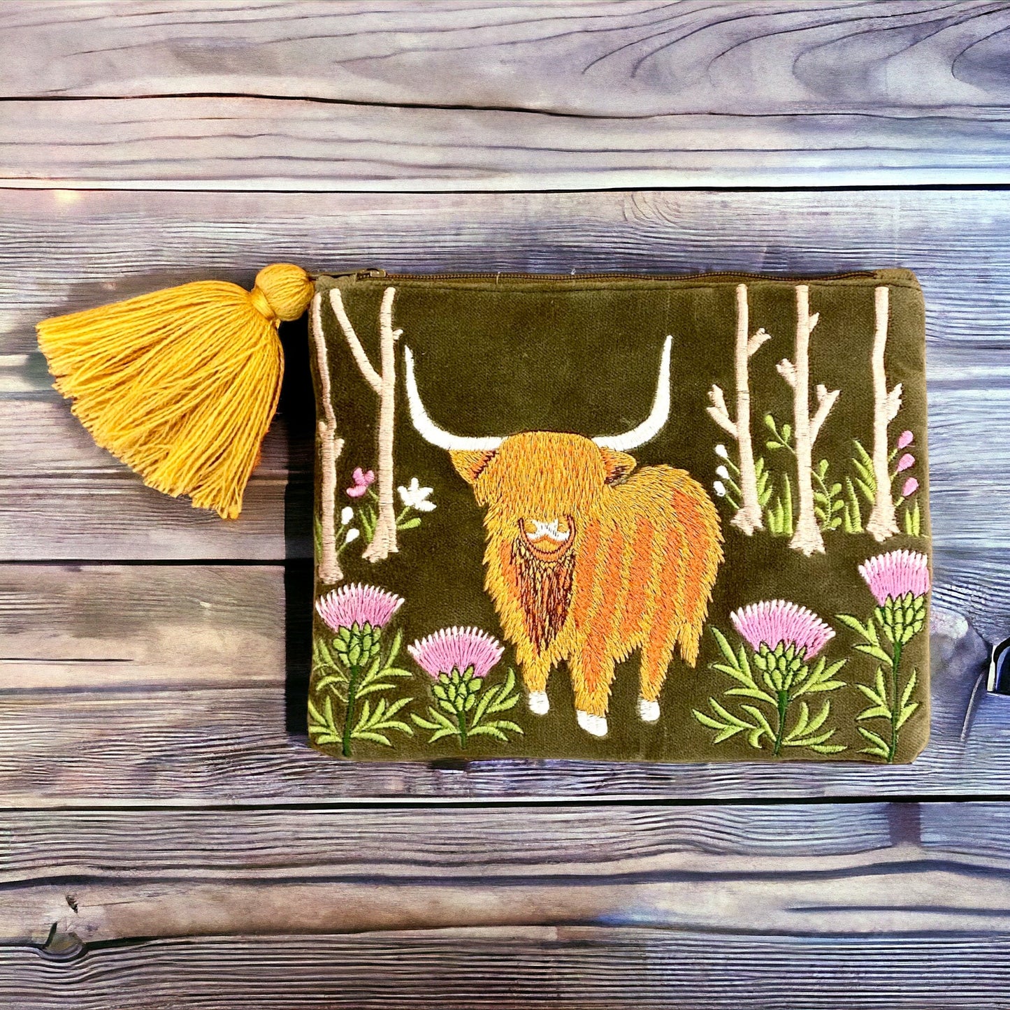 A green velvet pouch with an embroidered highland cow design, accompanied by a yellow tassel, placed on a wooden surface.