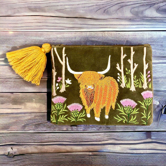 A green velvet pouch with an embroidered highland cow design, accompanied by a yellow tassel, placed on a wooden surface.