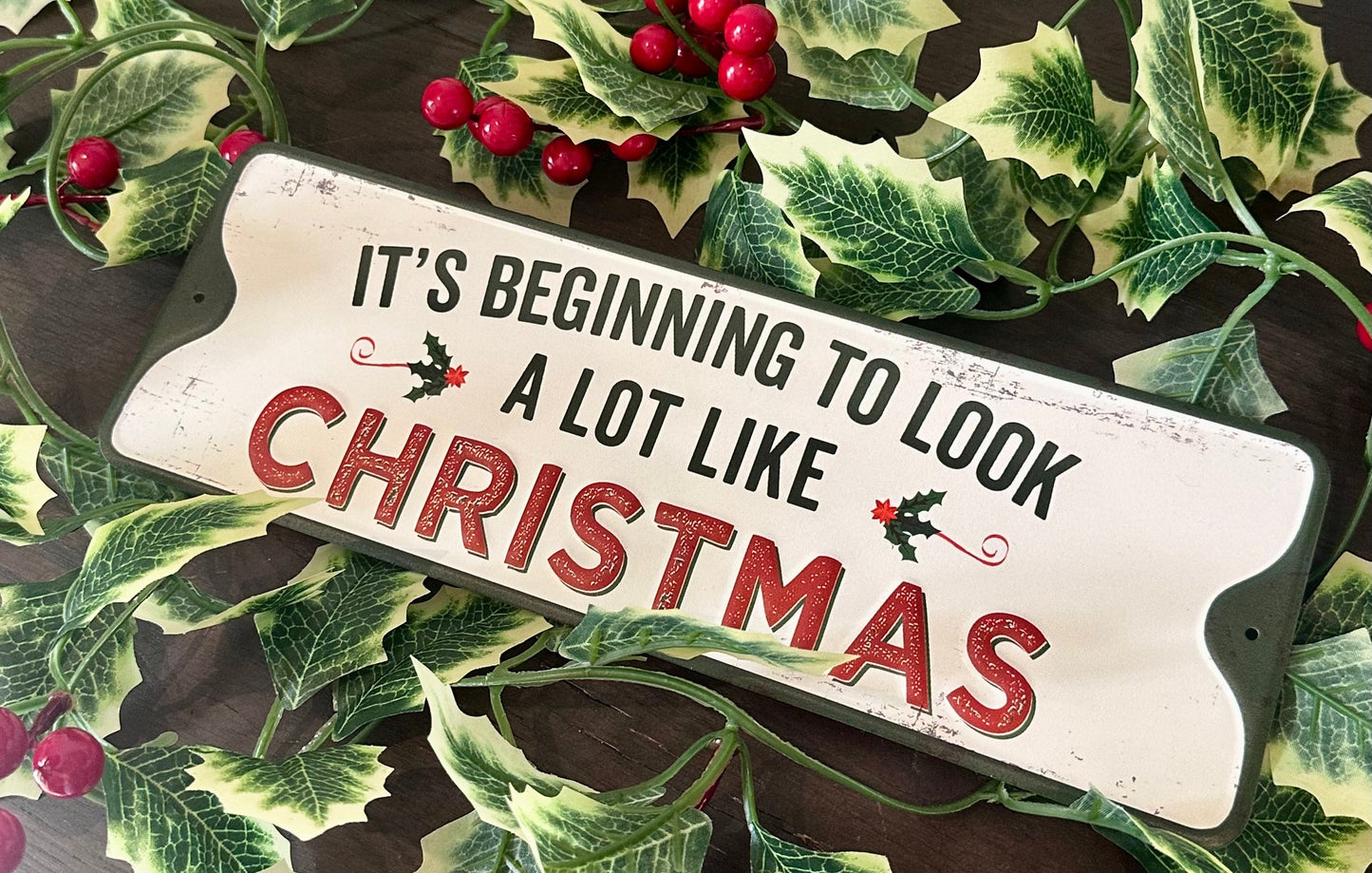A rustic white wooden Christmas sign with the text 'IT'S BEGINNING TO LOOK A LOT LIKE CHRISTMAS' in black and red lettering, surrounded by green holly leaves and red berries on a dark wooden surface.