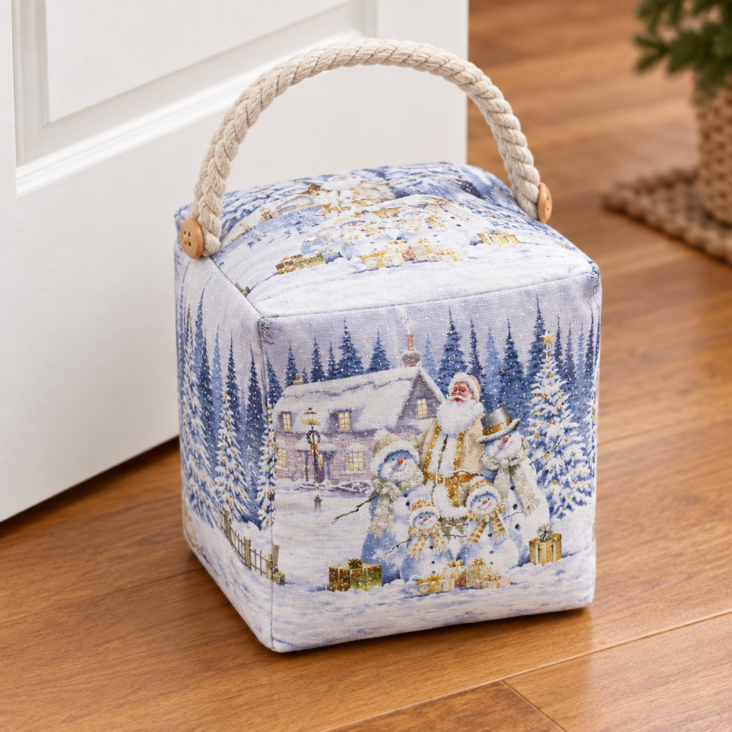 A square fabric doorstop with a rope handle, featuring a winter Christmas scene print with Santa, snowmen, and a snowy house. An inset in the top-left shows the bag illuminated with warm lights.