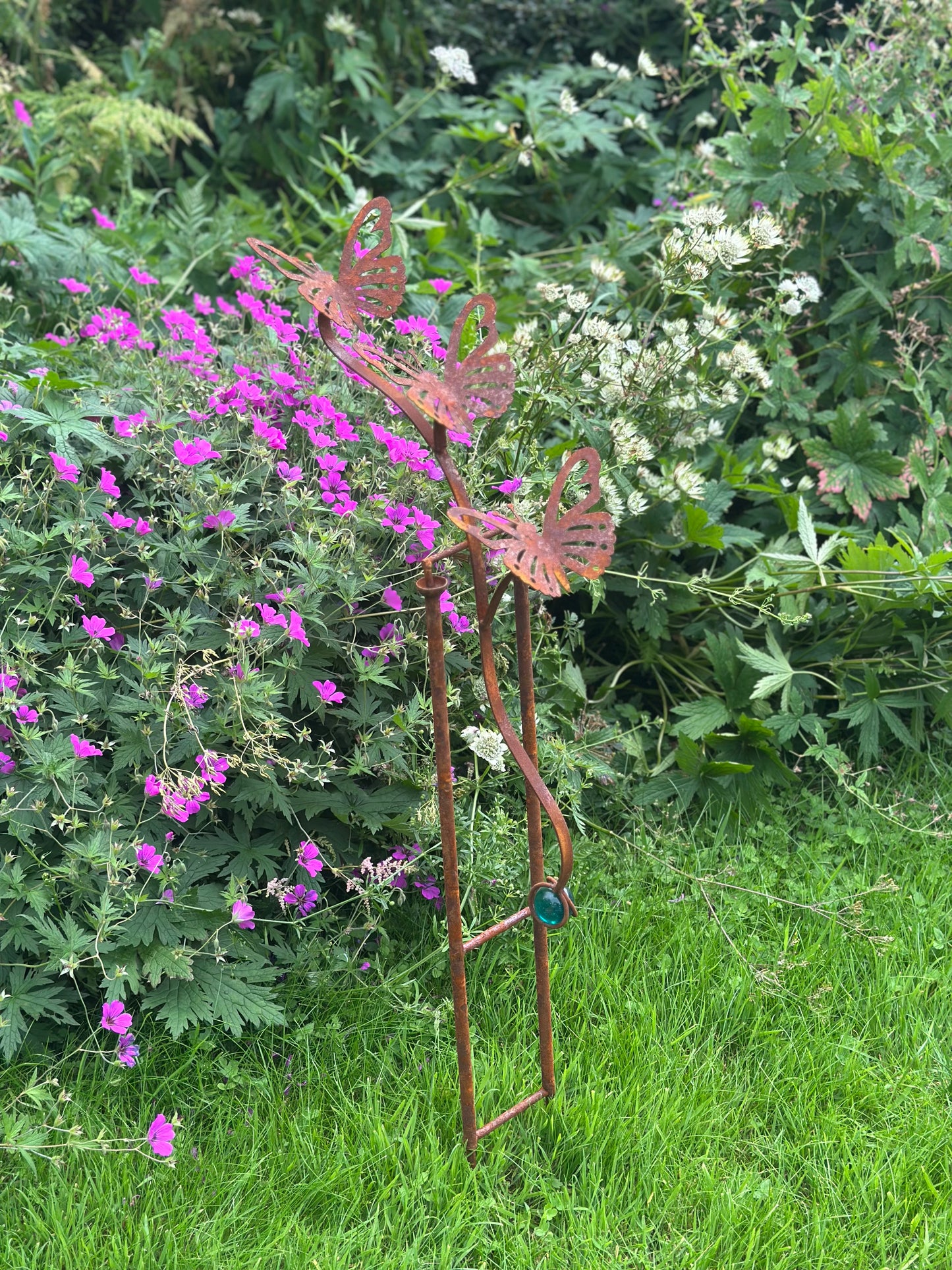 A rusted metal garden stake sculpture shaped like butterflies perched on a ladder, placed in a grassy garden bed with purple flowers and green foliage in the background.