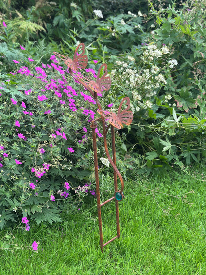 A rusted metal garden stake sculpture shaped like butterflies perched on a ladder, placed in a grassy garden bed with purple flowers and green foliage in the background.