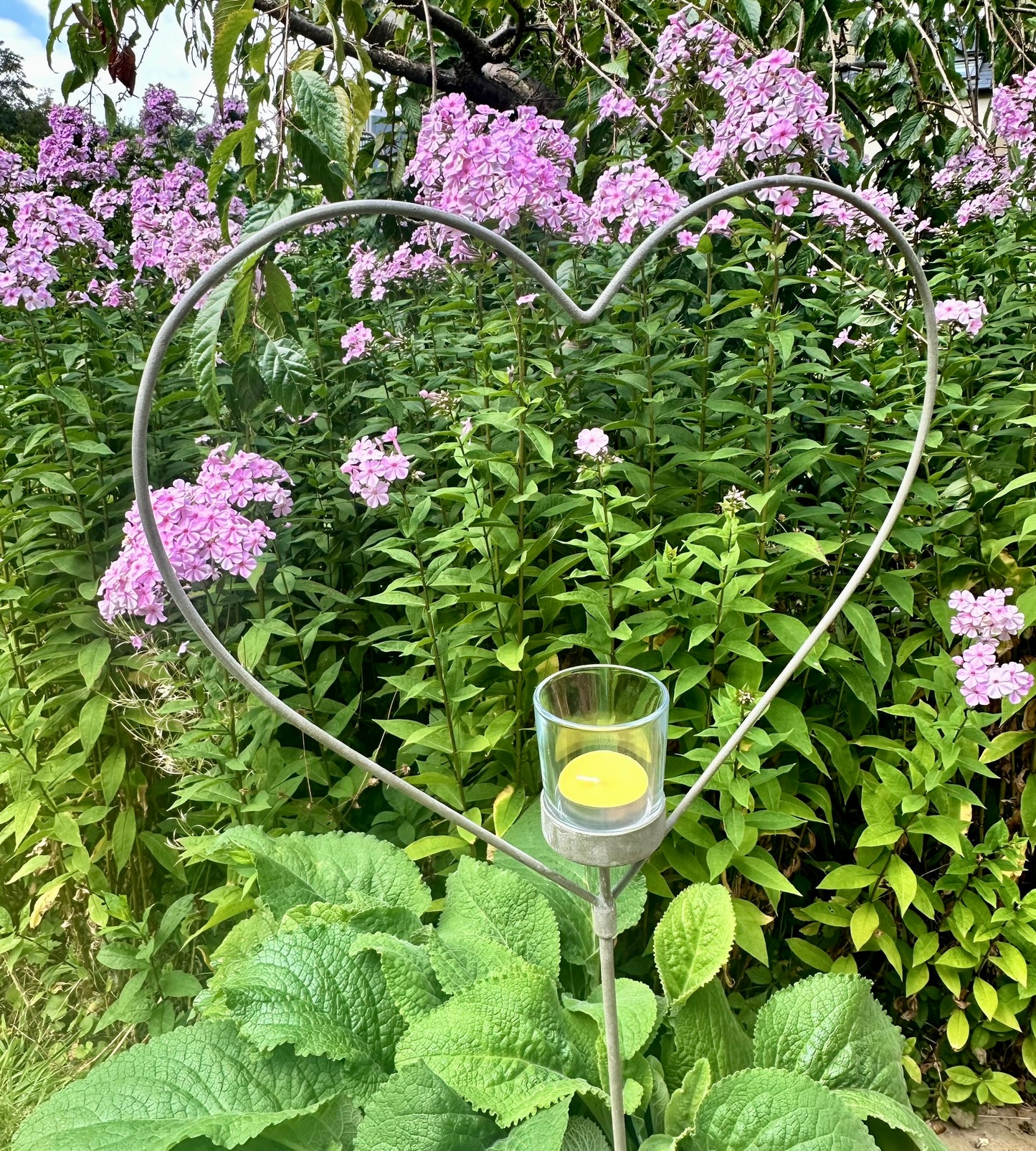 A silver-colored metal heart-shaped garden stake with a tealight candle placed in the center, set against a background of green foliage and purple flowers.