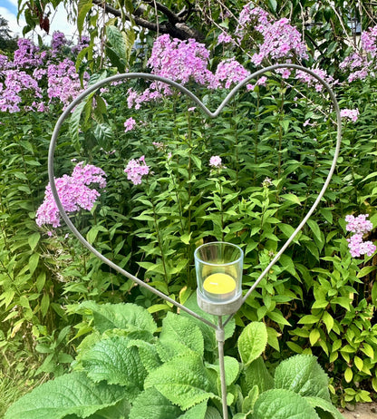 A silver-colored metal heart-shaped garden stake with a tealight candle placed in the center, set against a background of green foliage and purple flowers.