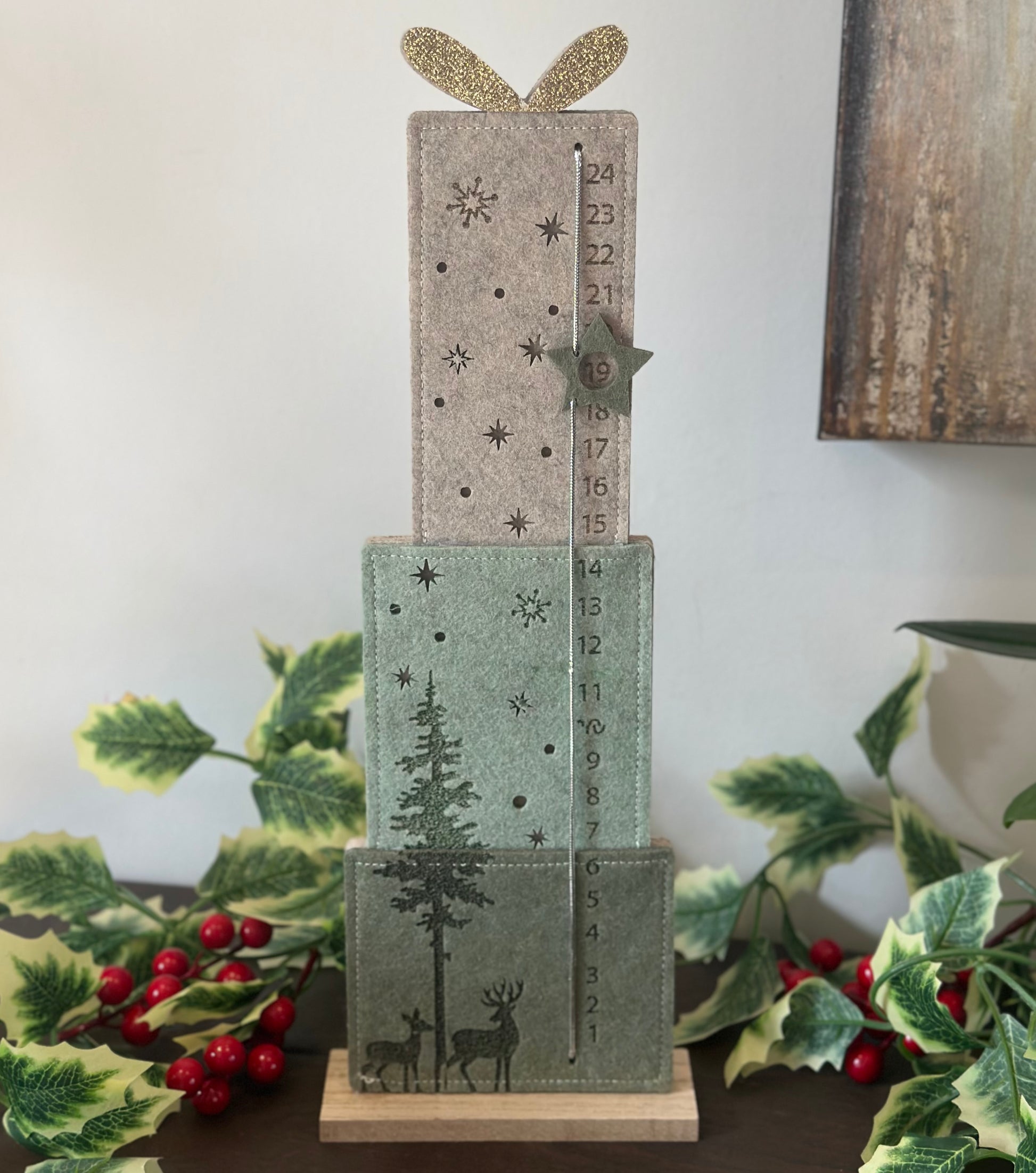 A three-tiered wooden advent calendar styled with green and beige felt panels decorated with Christmas motifs like trees, stars, reindeer, and numbers. The top tier has a glittery gold bow, and the calendar is displayed on a dark table with holiday greenery and red berries.