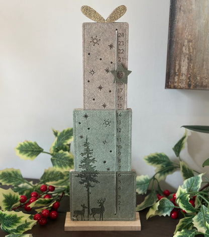 A three-tiered wooden advent calendar styled with green and beige felt panels decorated with Christmas motifs like trees, stars, reindeer, and numbers. The top tier has a glittery gold bow, and the calendar is displayed on a dark table with holiday greenery and red berries.