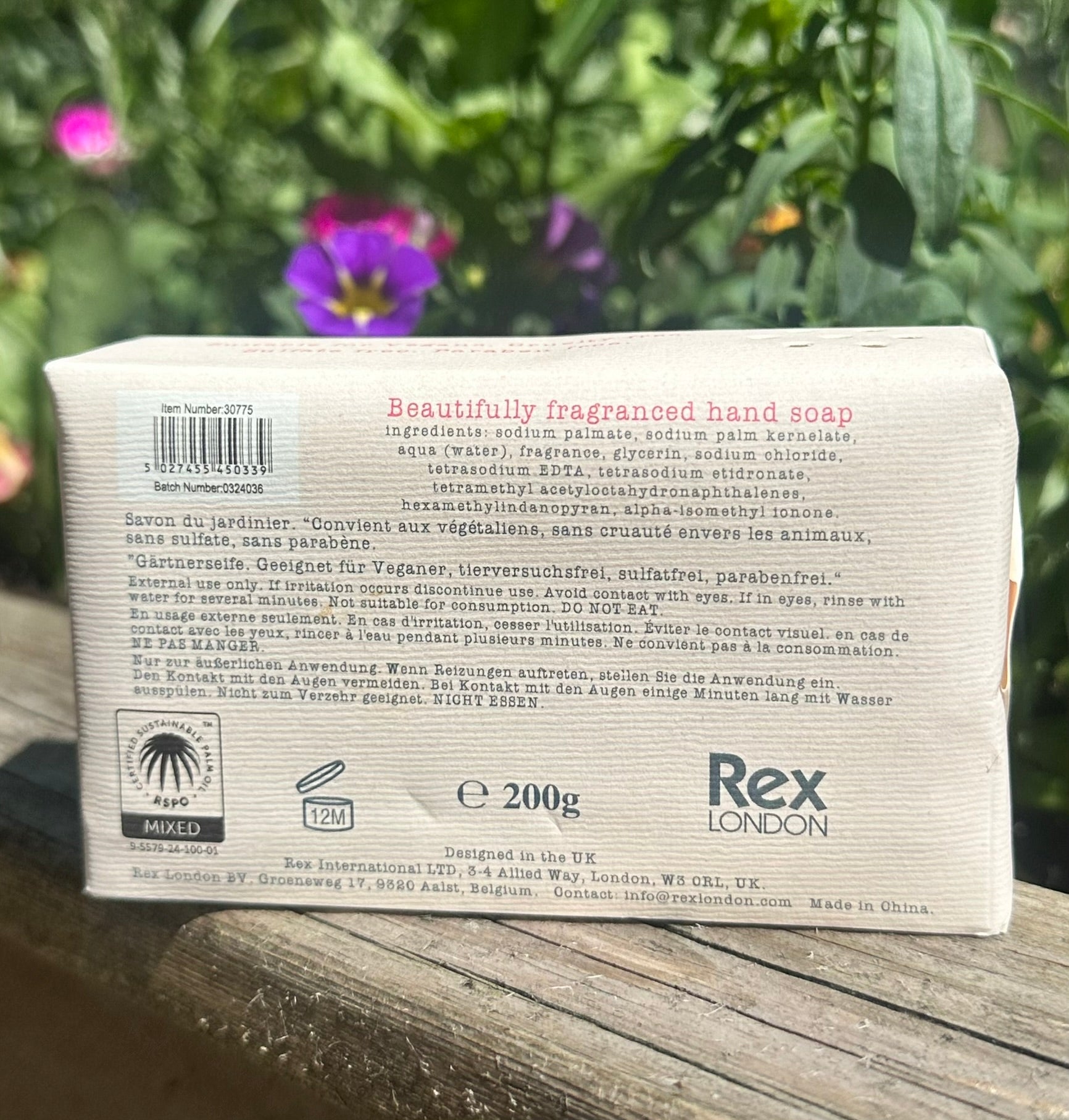 Back of a Rex London soap bar packaging with text and branding, placed on a wooden surface with greenery in the background.