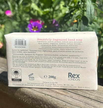 Back of a Rex London soap bar packaging with text and branding, placed on a wooden surface with greenery in the background.