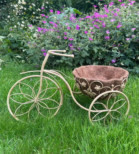 A decorative metal garden bench shaped like a vintage bicycle, with a woven brown planter basket on the back, placed on green grass in front of a flower bed.