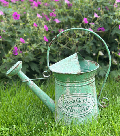 A distressed light green metal watering can with a conical spout and a handle loop, featuring a vintage-style label that reads 'Fresh Garden Flowers'. It is placed on green grass with a background of purple flowers.