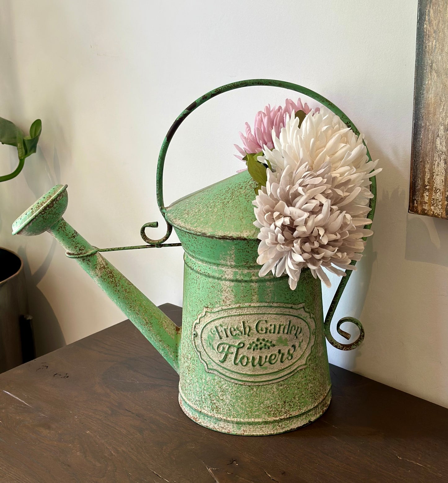 A distressed green metal watering can with a handle and spout, decorated with a vintage-style label reading 'Fresh Garden Flowers' and holding two large faux flowers, set on a dark wooden surface against a light wall.