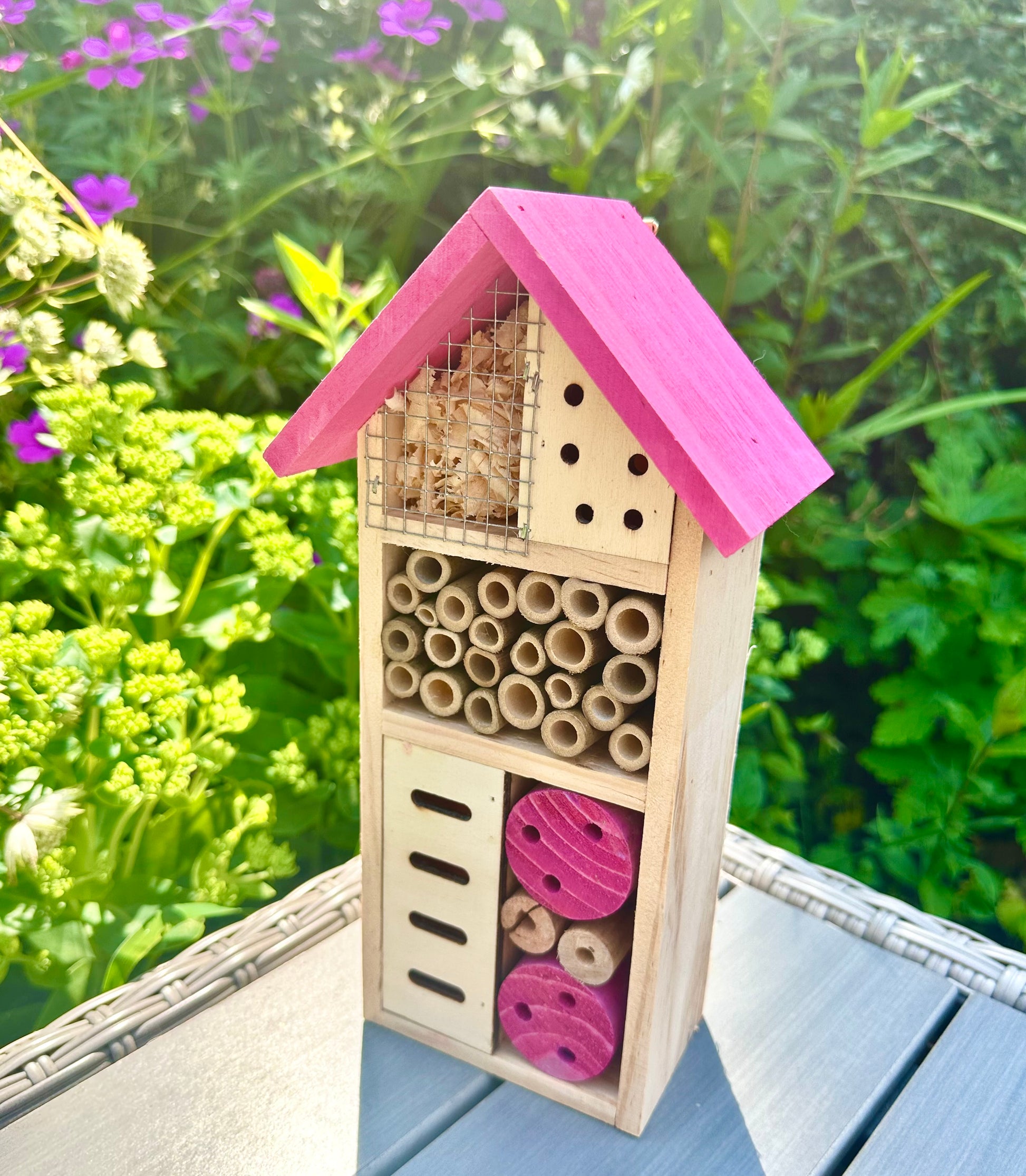 A wooden insect hotel with a pink roof and natural fiber inserts for different types of insects, displayed against a garden backdrop.